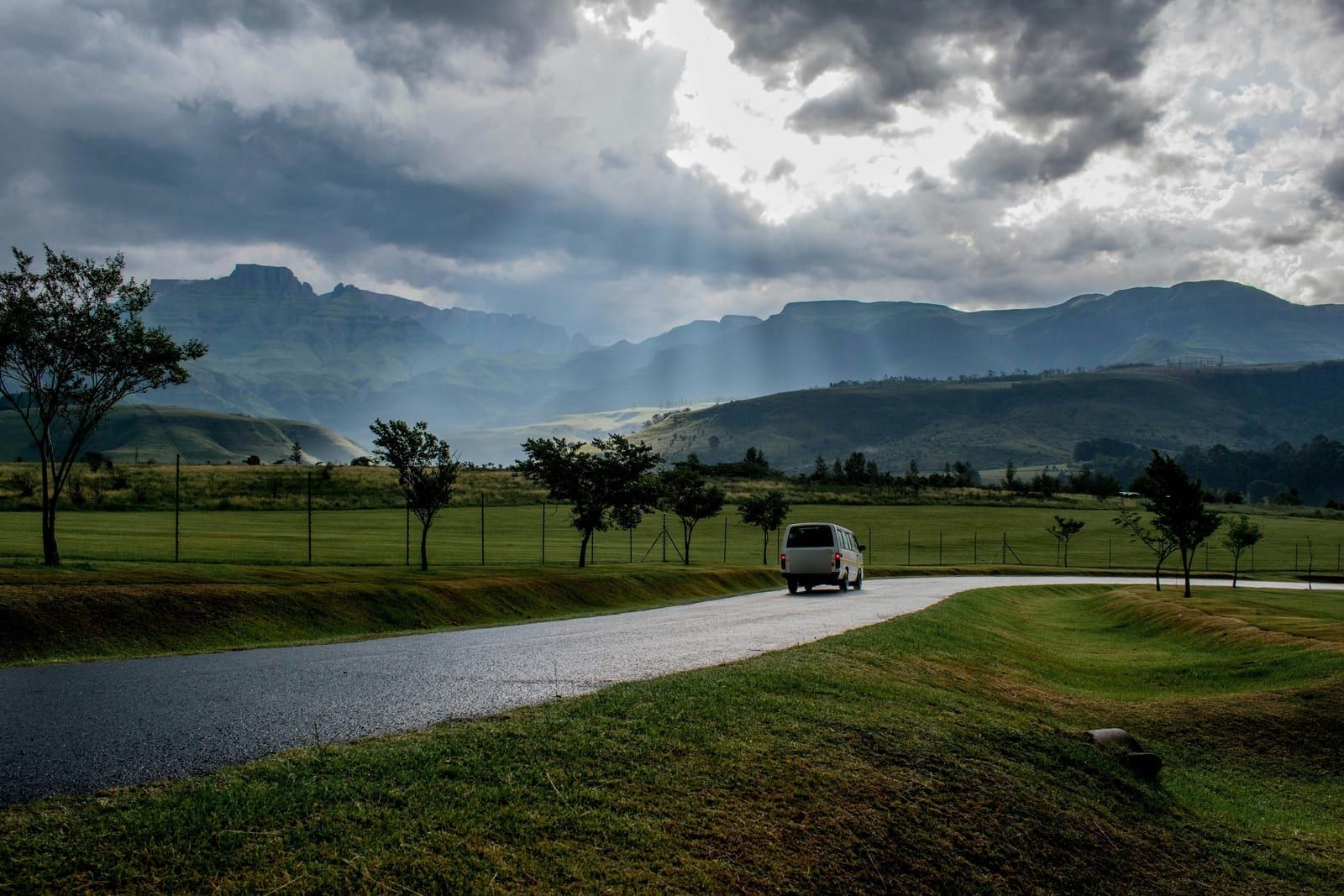 Beli kombi na mokrom putu kroz zelenu dolinu, ispod planina i oblaka